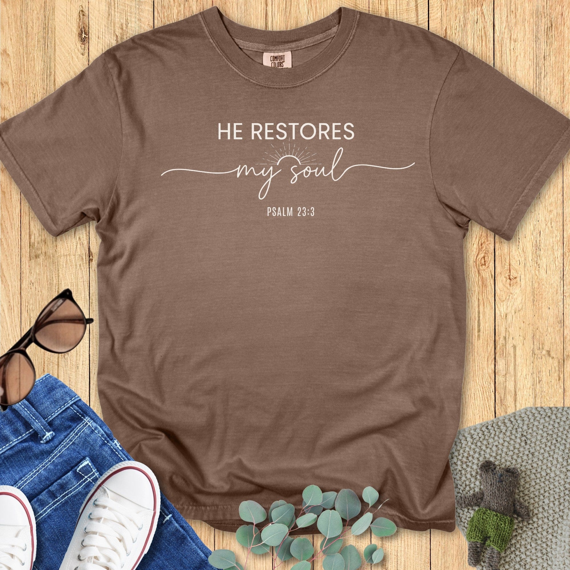 Comfort Colors 1717 espresso “He Restores My Soul – Psalm 23:3” t-shirt with minimalist sunrise graphic on rustic wood background. A cozy women’s scripture tee — meaningful faith-based apparel for women and modern Christian shirt, thoughtful Christian mom gift idea.
