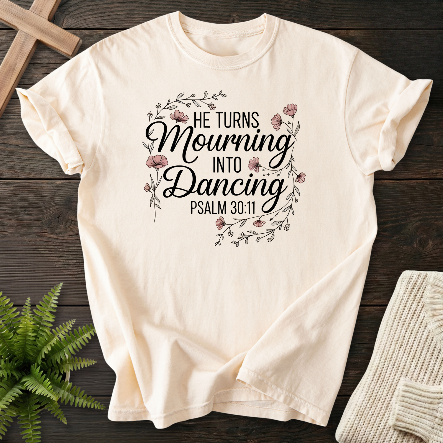 Ivory women's Christian t-shirt with a decorative flower wreath and "He Turns Mourning Into Dancing" script