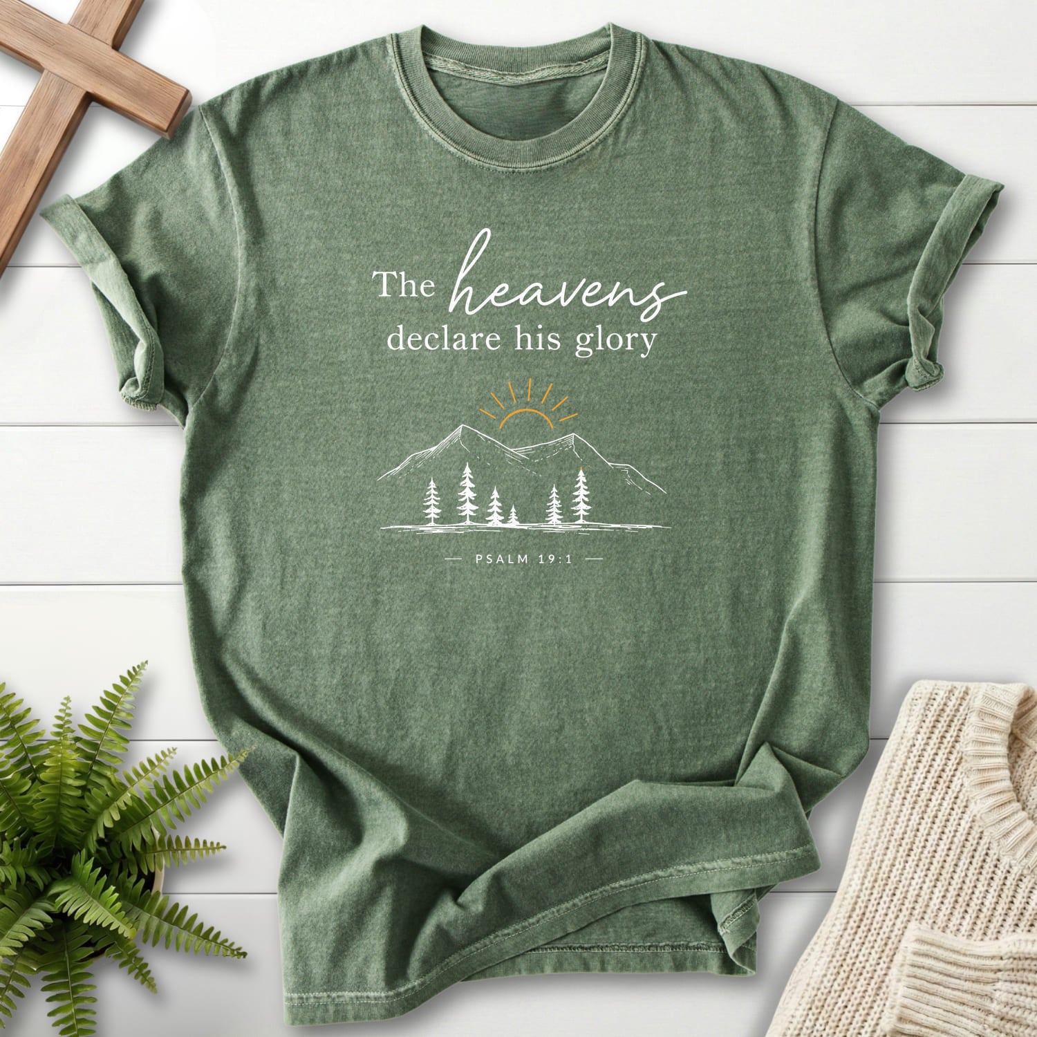 Flat lay of a Moss faith-based tee featuring sunrise graphic with "Heavens Declare His Glory Dark T-Shirt" Psalm 19:1.