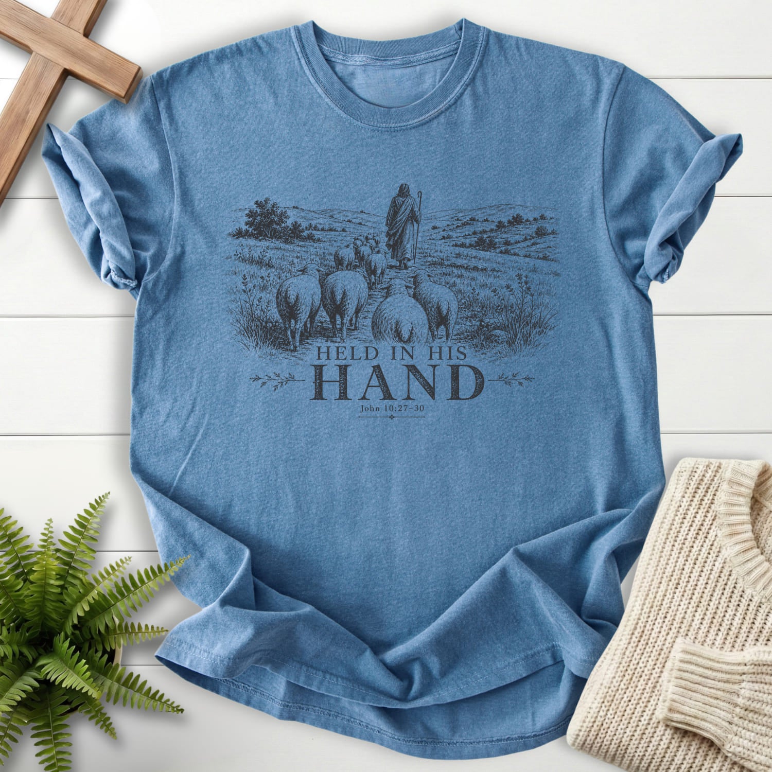 Flatlay of a Blue Jean Bible verse tee with "Held in His Hand T-Shirt" John 10:27-30.