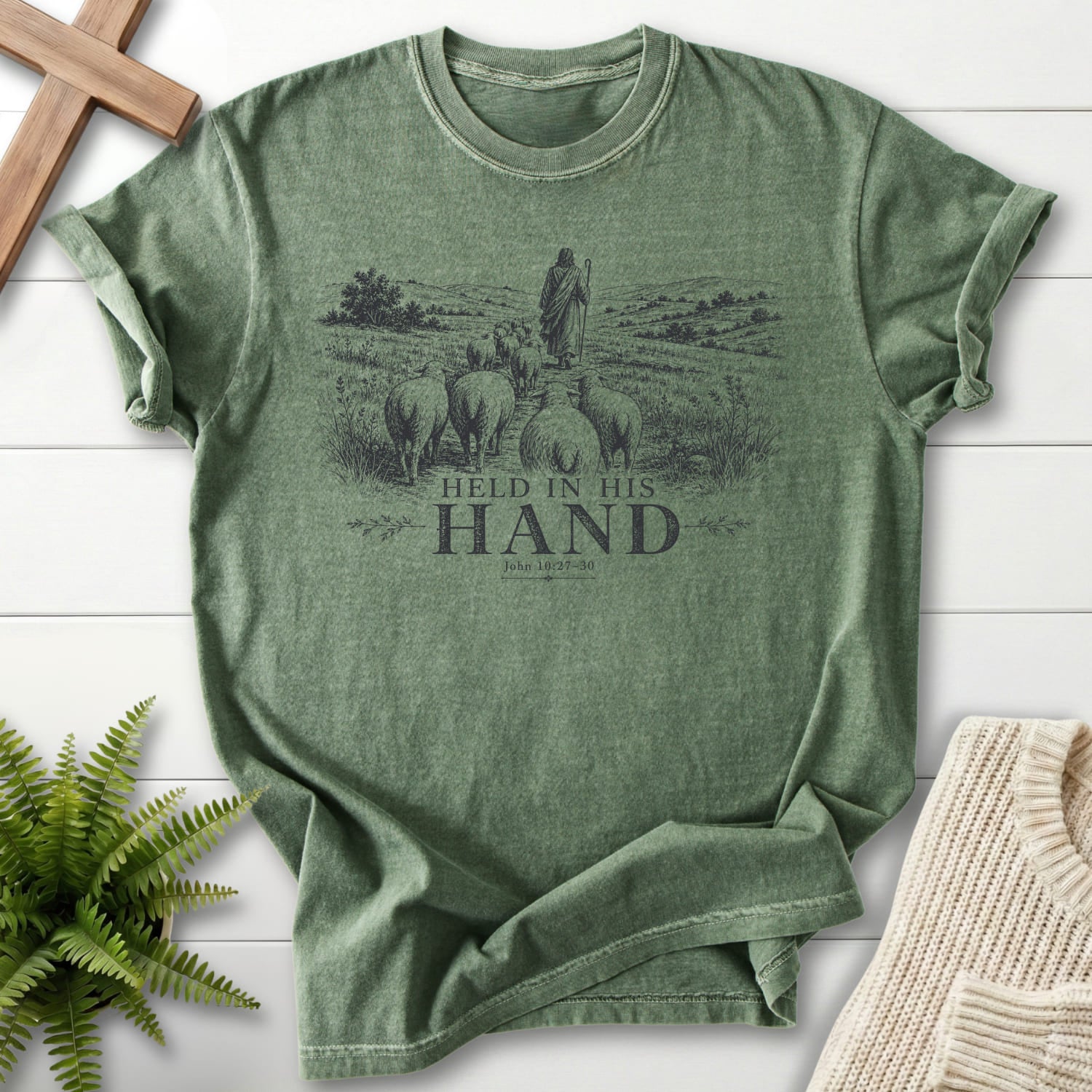 Flatlay of a Moss Christian graphic tee with "Held in His Hand T-Shirt" John 10:27-30.