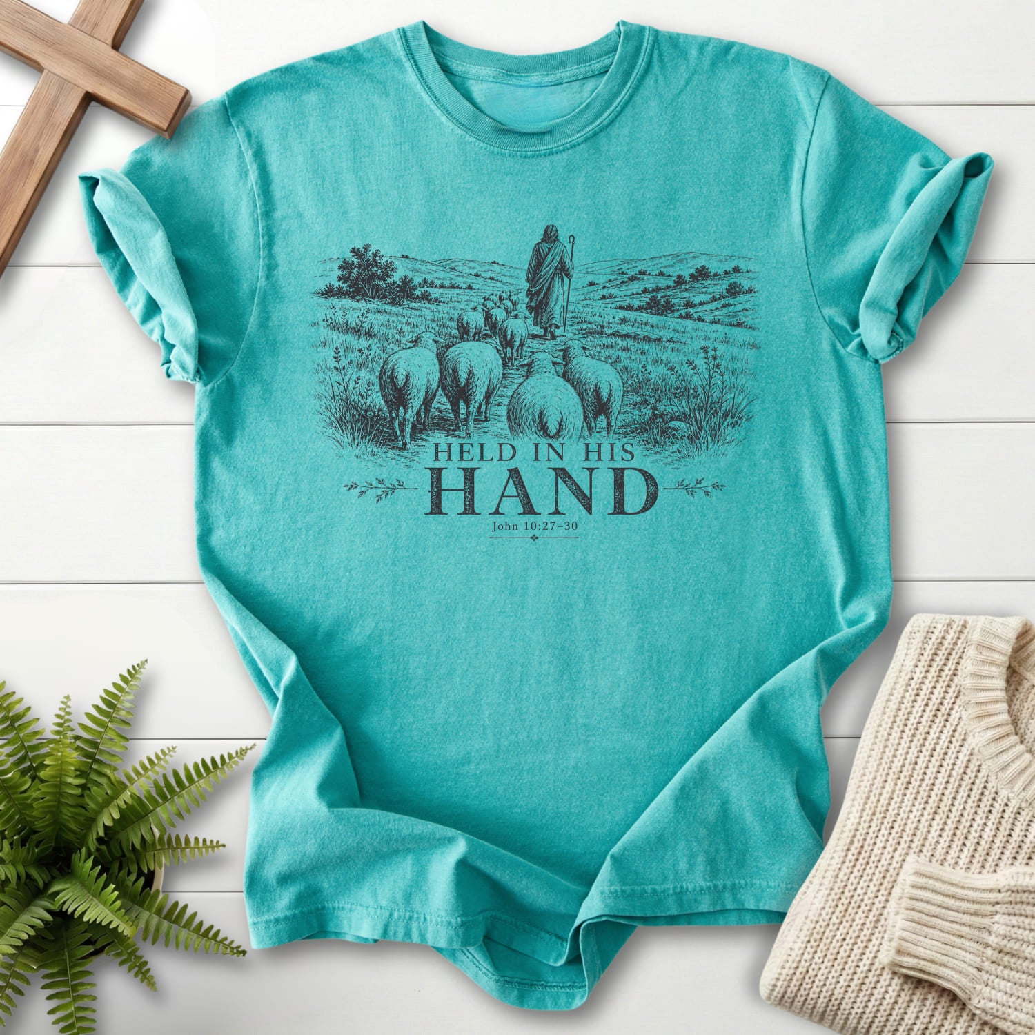 Flat lay of a Seafoam faith-inspired shirt featuring "Held in His Hand T-Shirt" John 10:27-30.