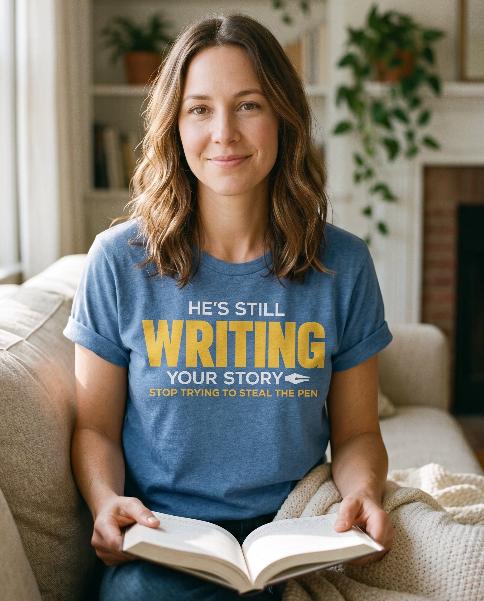 He's Still Writing Your Story – Blue Jean women's Christian tee, lifestyle photo in cozy living room – Bold lettering "He's Still Writing Your Story – She Wears Faith