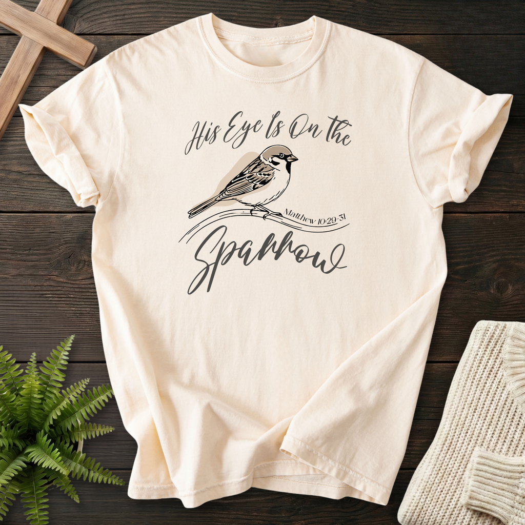 Ivory women's Christian t-shirt with perched bird artwork and flowing "His Eye Is on the Sparrow" script, inspired by Matthew 10:29-31