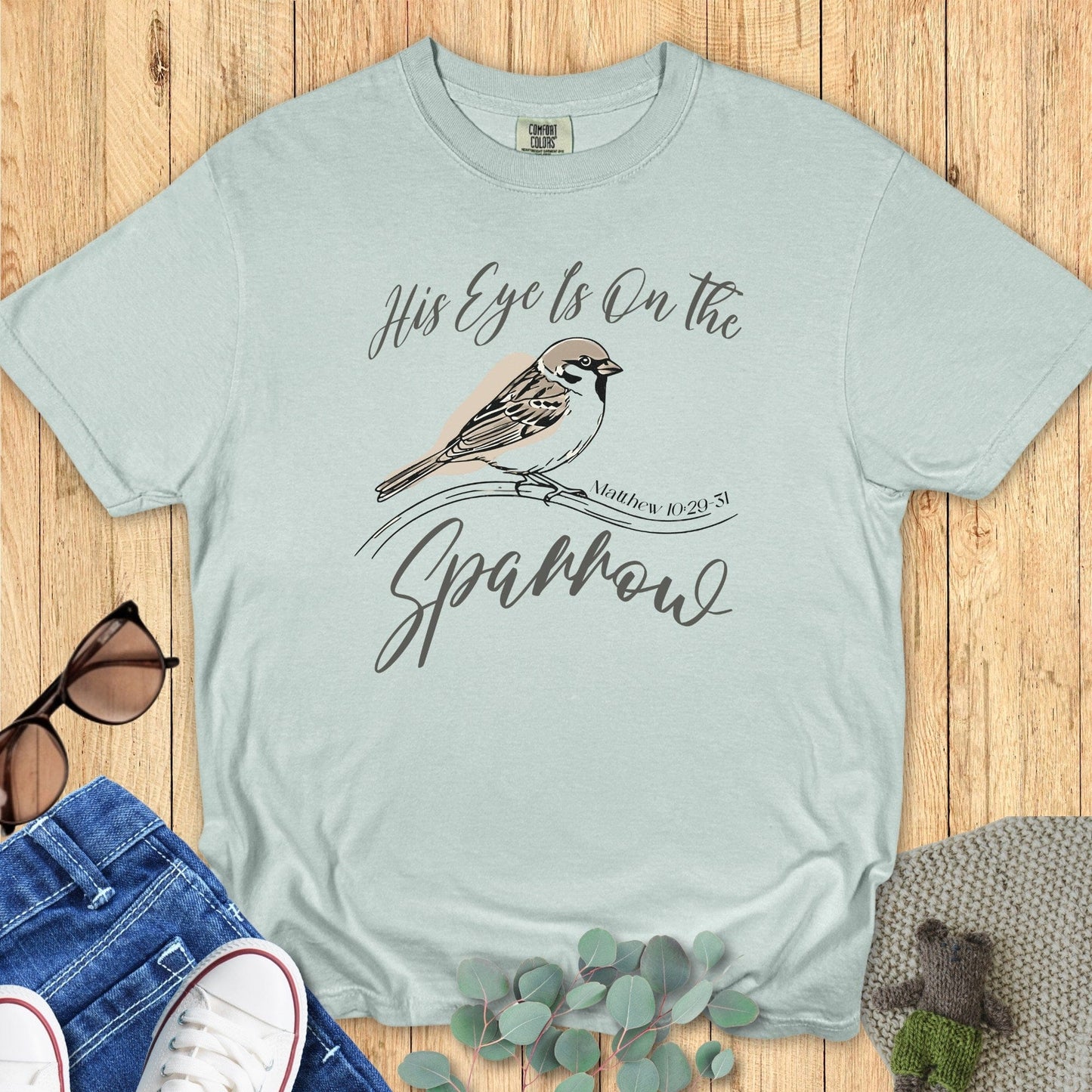 Flatlay of a Comfort Colors 1717 bay “His Eye Is on the Sparrow – Matthew 10:29-31” t-shirt featuring a detailed bird design and elegant script typography. A serene women’s scripture tee — inspiring faith-based apparel for women and modern Christian shirt, perfect Christian mom gift idea.