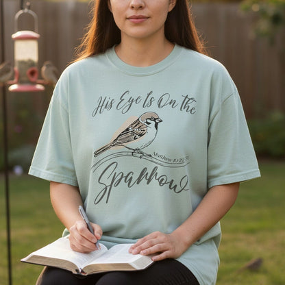 Woman journaling outdoors while wearing a Comfort Colors 1717 bay “His Eye Is on the Sparrow – Matthew 10:29-31” t-shirt featuring a hand-drawn sparrow on a branch. A peaceful women’s scripture tee — encouraging faith-based apparel for women and modern Christian shirt, thoughtful Christian mom gift idea.
