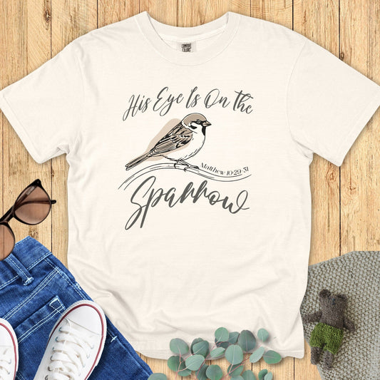 Flatlay of a Comfort Colors 1717 ivory “His Eye Is on the Sparrow – Matthew 10:29-31” t-shirt with delicate sparrow illustration and scripture text. A soft women’s scripture tee — uplifting faith-based apparel for women and modern Christian shirt, ideal Christian mom gift idea.