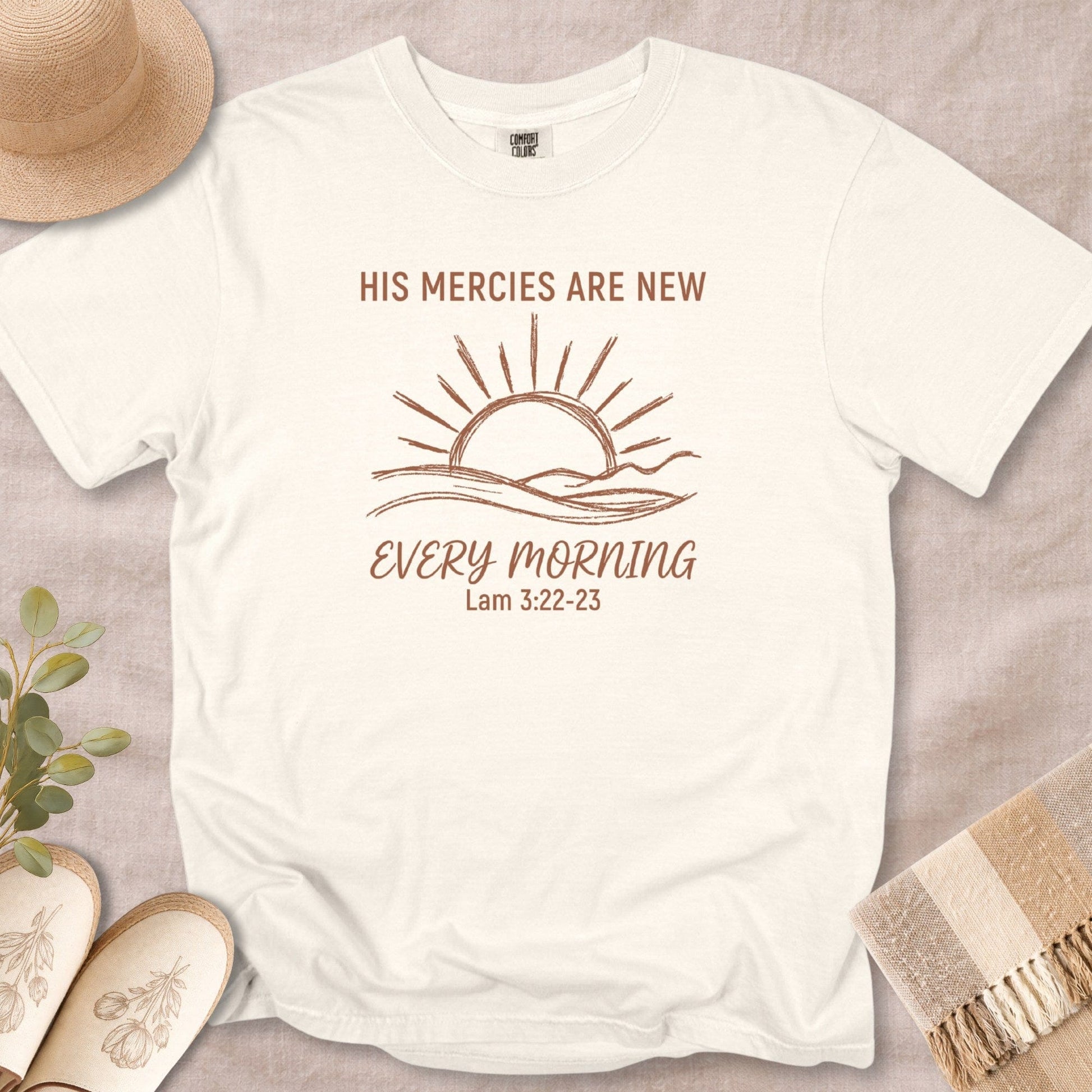 Flat lay of an Ivory Comfort Colors 1717 tshirt with a sun and waves illustration. A spiritual Christian women tee with the Bible verse "His Mercies Are New Every Morning," designed for US faith wear collections.