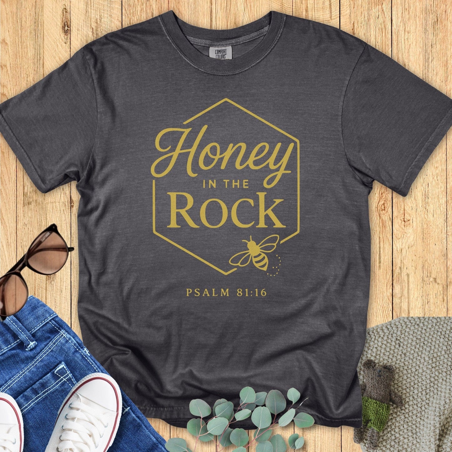 Flatlay of a Comfort Colors 1717 pepper “Honey in the Rock – Psalm 81:16” t-shirt featuring a gold honeycomb frame and bee illustration. A timeless women’s scripture tee — modern Christian shirt and elegant faith-based apparel for women, perfect Christian mom gift idea.
