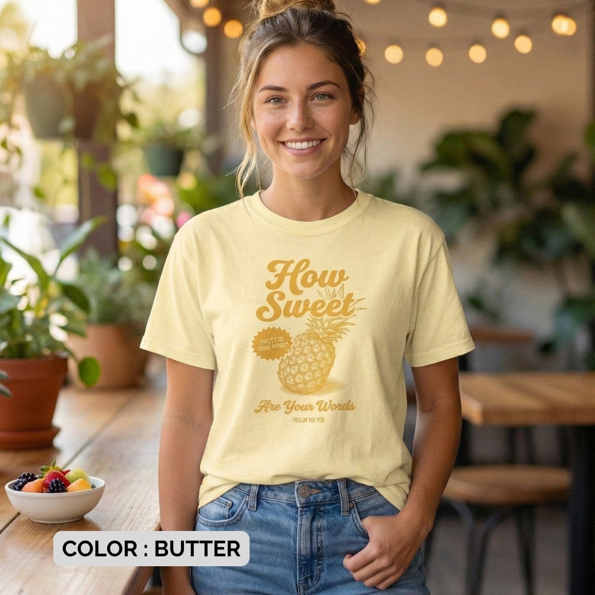 Christian women tee in butter Comfort Colors 1717 featuring the “How Sweet Are Your Words” Psalm 119:103 pineapple design, worn by a smiling woman in a cozy café setting with warm lights and plants; inspirational US faith wear.
