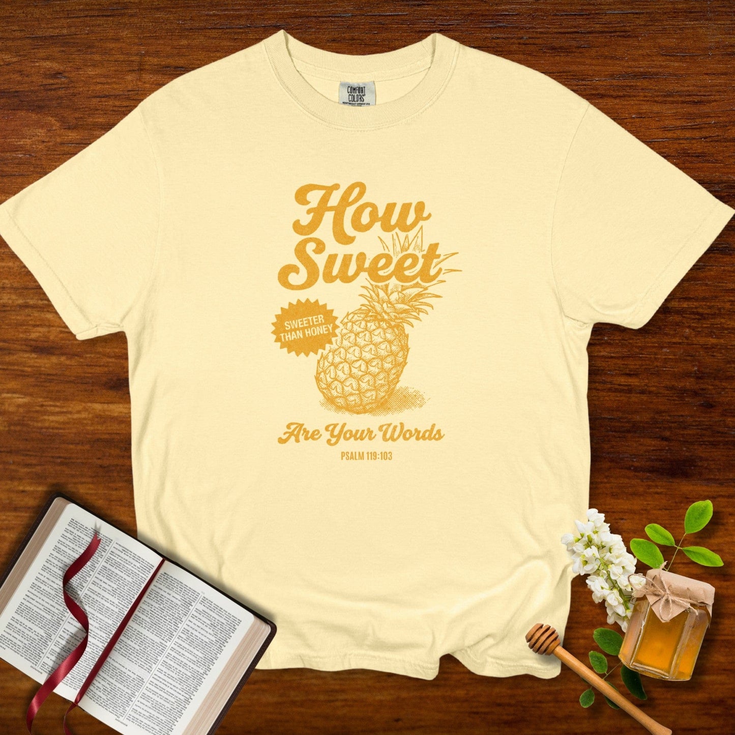 Christian women tee featuring a “How Sweet Are Your Words” Psalm 119:103 pineapple design printed on a soft butter Comfort Colors 1717 shirt, styled with an open Bible and honey jar on a wooden background; US faith wear apparel.