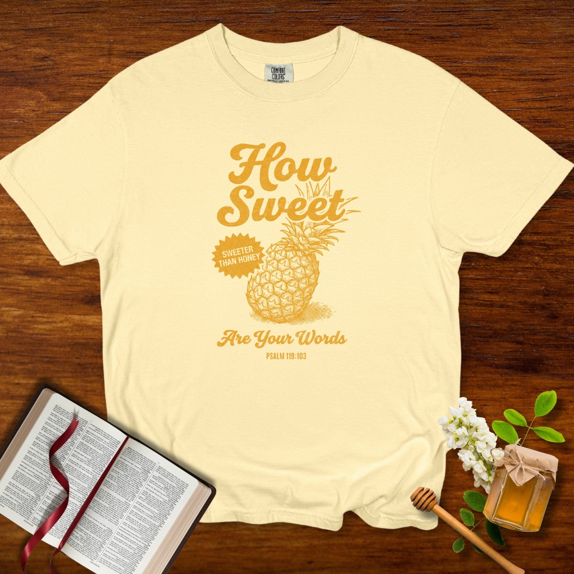 Christian women tee featuring a “How Sweet Are Your Words” Psalm 119:103 pineapple design printed on a soft butter Comfort Colors 1717 shirt, styled with an open Bible and honey jar on a wooden background; US faith wear apparel.