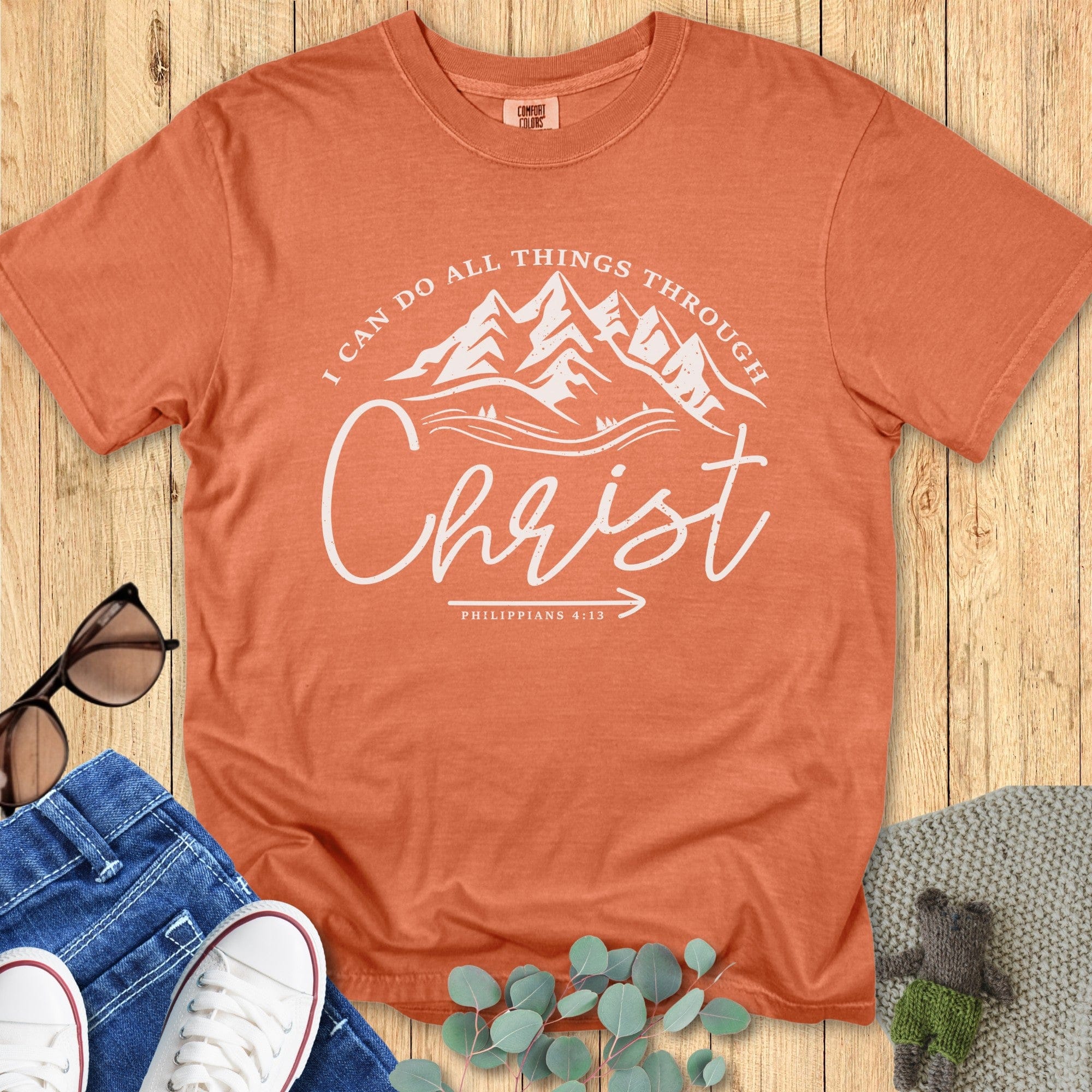 Flatlay of a Comfort Colors 1717 yam orange “I Can Do All Things Through Christ – Philippians 4:13” t-shirt with mountain illustration. A warm and inspiring faith-based apparel for women piece.