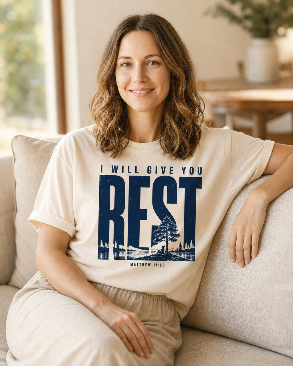 I Will Give You Rest – Ivory women's Christian tee, lifestyle photo in cozy living room – Mountain lake scene inside bold REST lettering – Matthew 11:28 – She Wears Faith