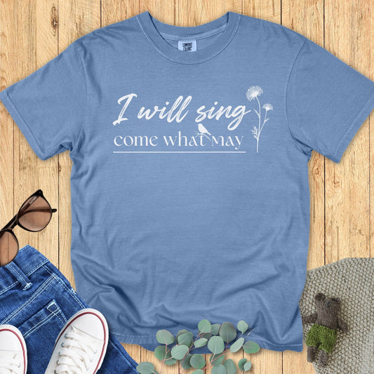 Flatlay of a Comfort Colors 1717 blue jean “I Will Sing Come What May” t-shirt with white script typography and botanical line art. A stylish women’s scripture tee — relaxed faith-based apparel for women and perfect modern Christian shirt, making a heartfelt Christian mom gift idea.