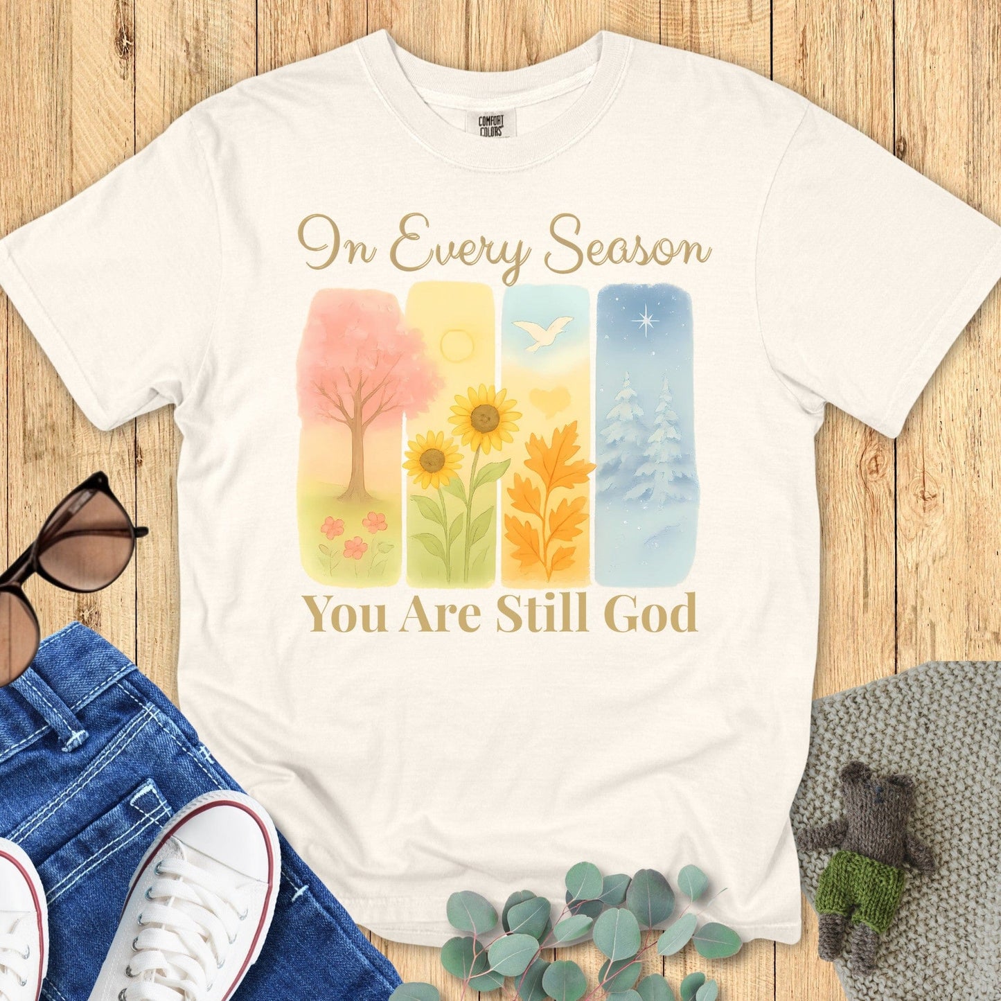 Flatlay of a Comfort Colors 1717 ivory “In Every Season You Are Still God” t-shirt with watercolor seasonal art and elegant script. A timeless women’s scripture tee and modern Christian shirt that celebrates God’s constant presence.