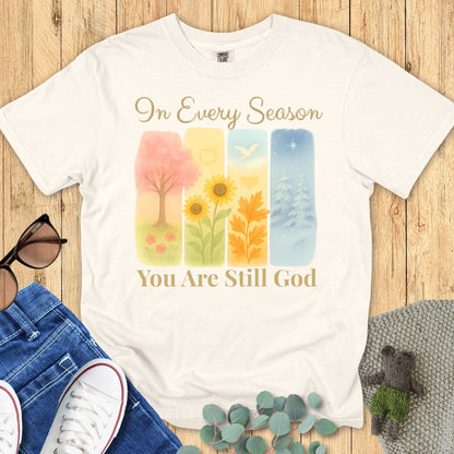 Flatlay of a Comfort Colors 1717 ivory “In Every Season You Are Still God” t-shirt with watercolor seasonal art and elegant script. A timeless women’s scripture tee and modern Christian shirt that celebrates God’s constant presence.