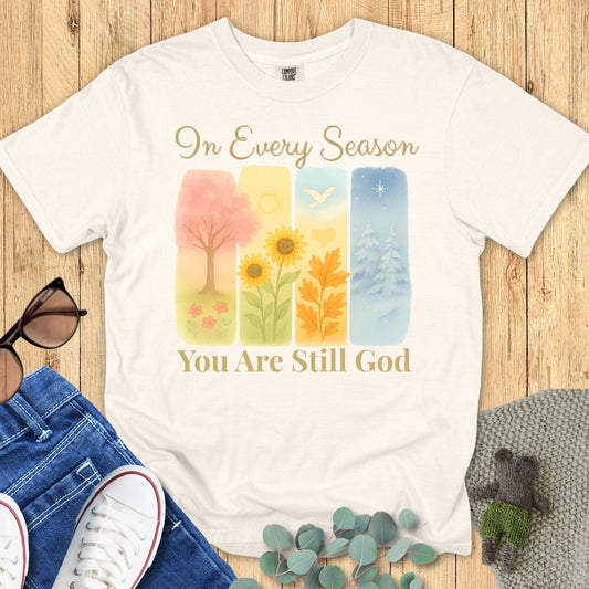 Flatlay of a Comfort Colors 1717 ivory “In Every Season You Are Still God” t-shirt with watercolor seasonal art and elegant script. A timeless women’s scripture tee and modern Christian shirt that celebrates God’s constant presence.