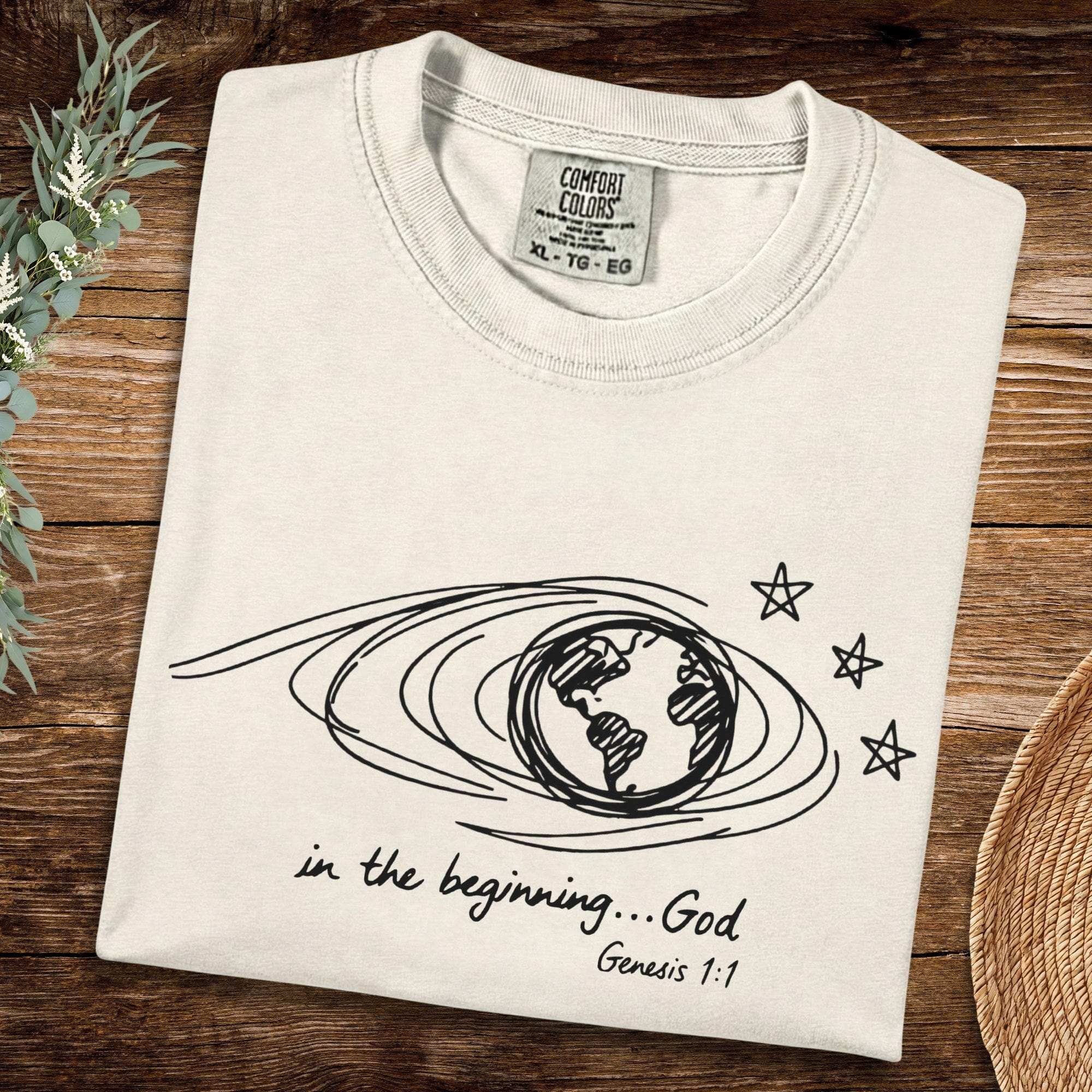 Flat lay of an Ivory Comfort Colors 1717 tshirt featuring black naive scribble art of the earth and stars. Minimalist Christian women tee with "In the beginning... God" Genesis 1:1 scripture, perfect for US faith wear collections.