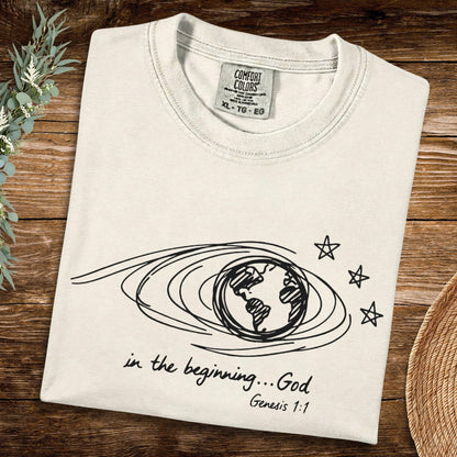 Flat lay of an Ivory Comfort Colors 1717 tshirt featuring black naive scribble art of the earth and stars. Minimalist Christian women tee with "In the beginning... God" Genesis 1:1 scripture, perfect for US faith wear collections.