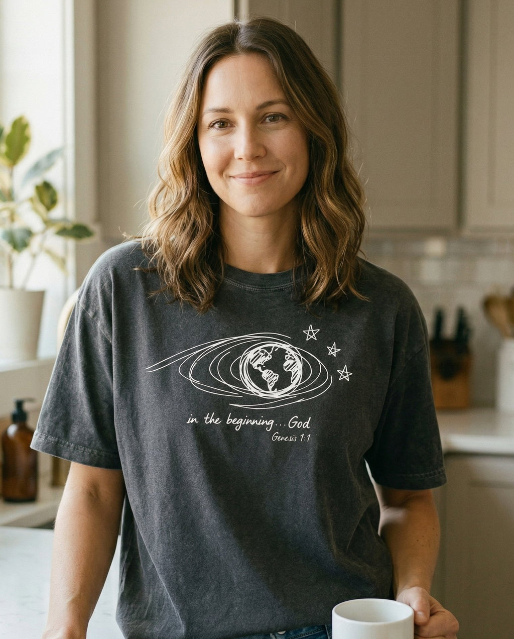 In the beginning God – Pepper women's Christian tee, lifestyle photo in morning kitchen setting – Minimalist line-art globe with orbital swirl and scattered stars with  – Genesis 1:1 – She Wears Faith