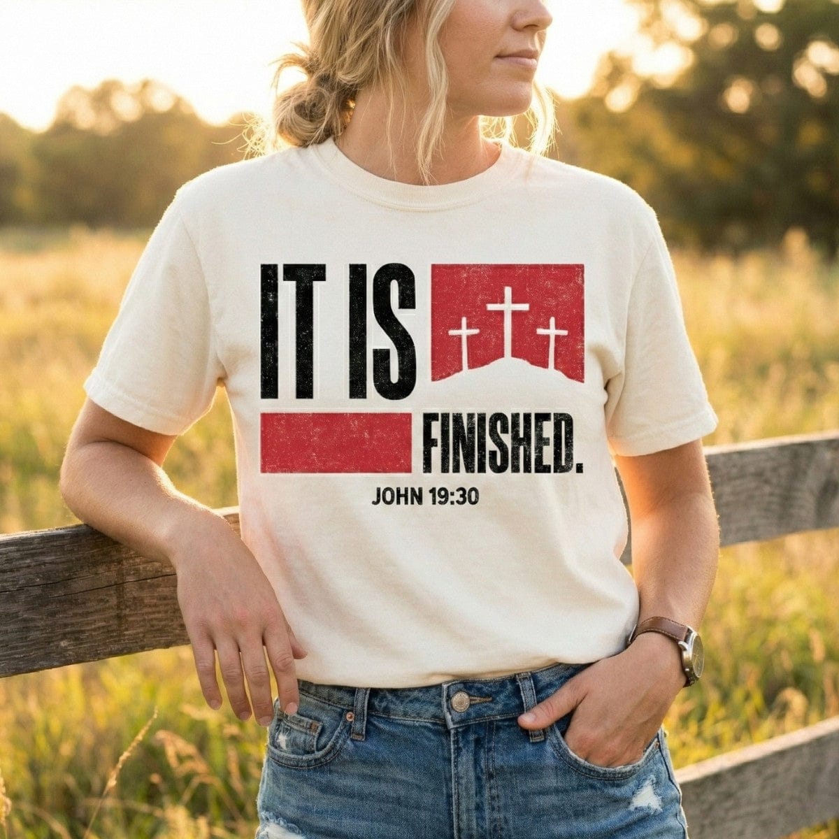 Woman leaning on a wooden fence wearing an Ivory Comfort Colors 1717 tshirt with a bold red and black "It Is Finished" design featuring three crosses. Lifestyle photography of a Christian women tee with John 19:30 scripture, styled for US faith wear collections.