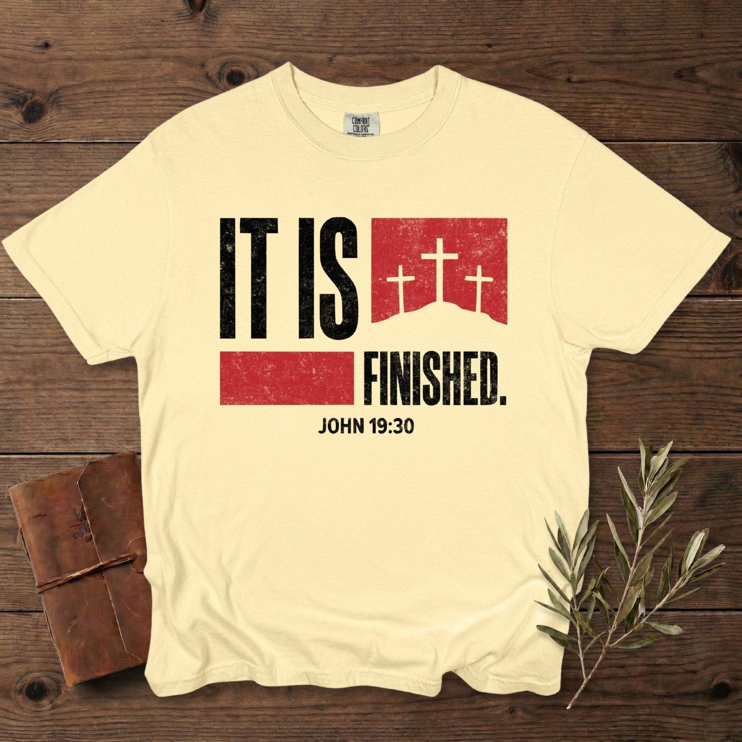 Comfort Colors 1717 butter yellow Christian women tshirt showcasing the “It Is Finished – John 19:30” scripture design with distressed black and red graphics and three crosses. Faith-based US faith wear tee styled on a dark wood background with a journal and olive branch.