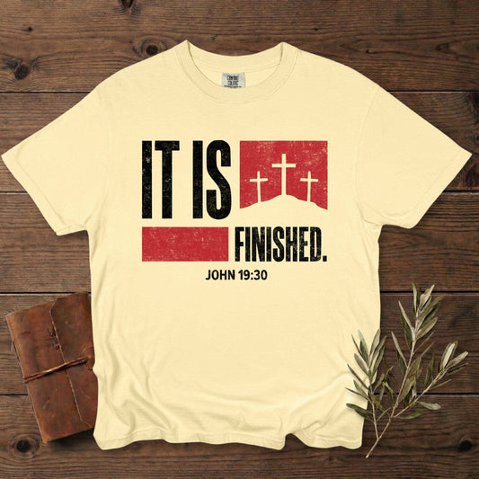 Comfort Colors 1717 butter yellow Christian women tshirt showcasing the “It Is Finished – John 19:30” scripture design with distressed black and red graphics and three crosses. Faith-based US faith wear tee styled on a dark wood background with a journal and olive branch.