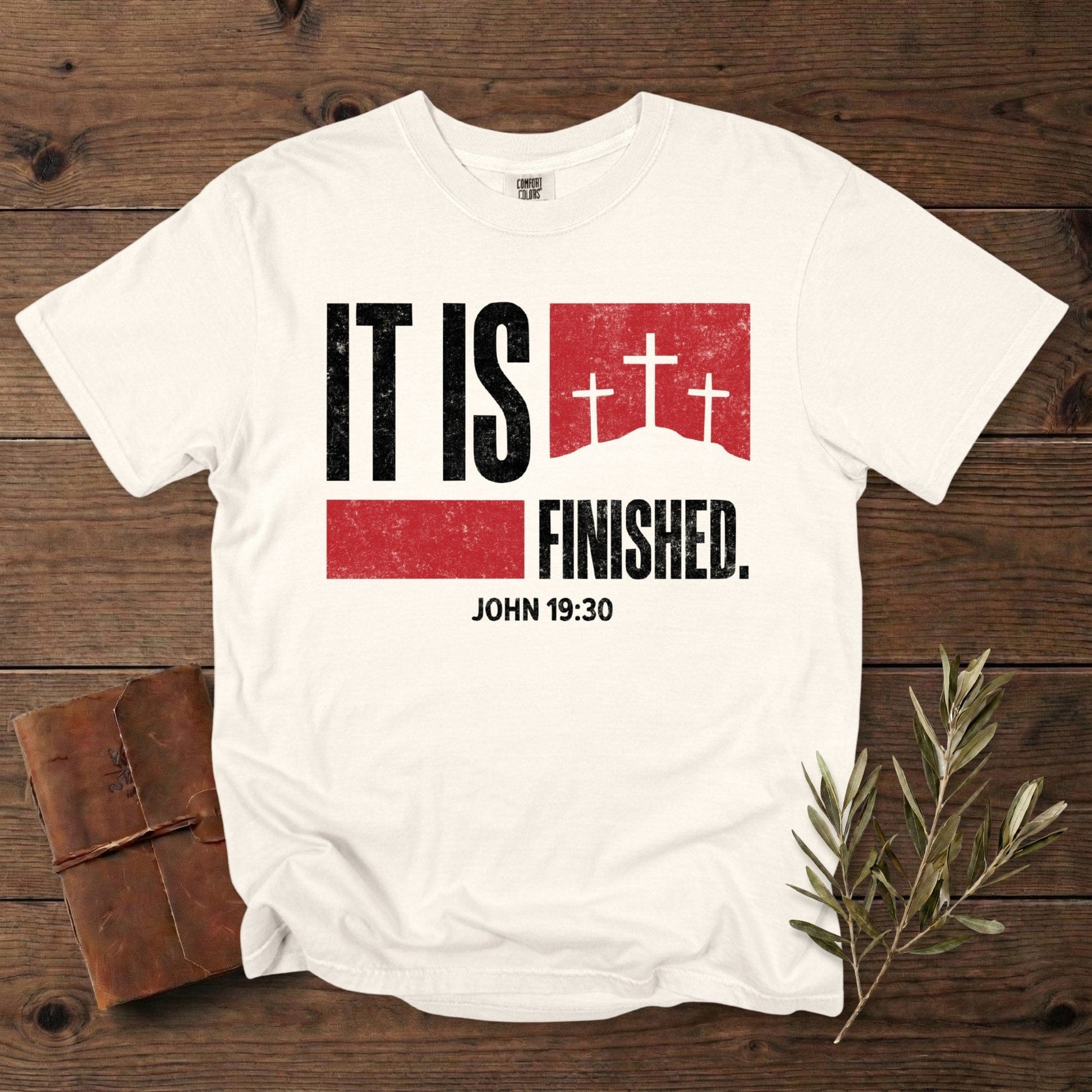 Comfort Colors 1717 ivory Christian women tee featuring the “It Is Finished – John 19:30” bold scripture design with red blocks and three cross silhouettes. Classic Christian tshirt for US faith wear, laid out on a rustic wooden surface with a journal and olive twig decor.