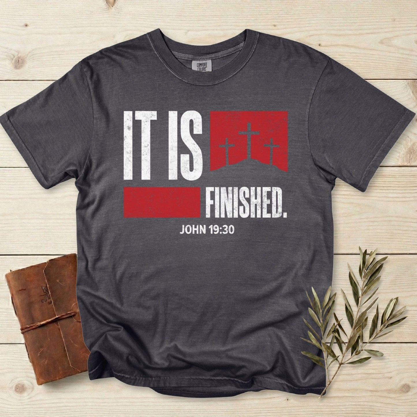 Comfort Colors 1717 pepper Christian women tee featuring a bold “It Is Finished – John 19:30” design with red distressed blocks and three crosses on Calvary. Inspirational Christian tshirt for US faith wear, displayed on a light wood background with a leather journal and olive branch.