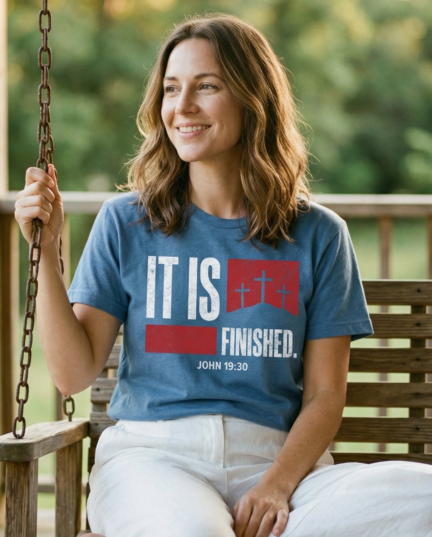 It Is Finished – Blue Jean women's Christian tee, lifestyle photo in sunny porch – Bold grunge-style typography reading 'It Is Finished' with a red banne – John 19:30 – She Wears Faith