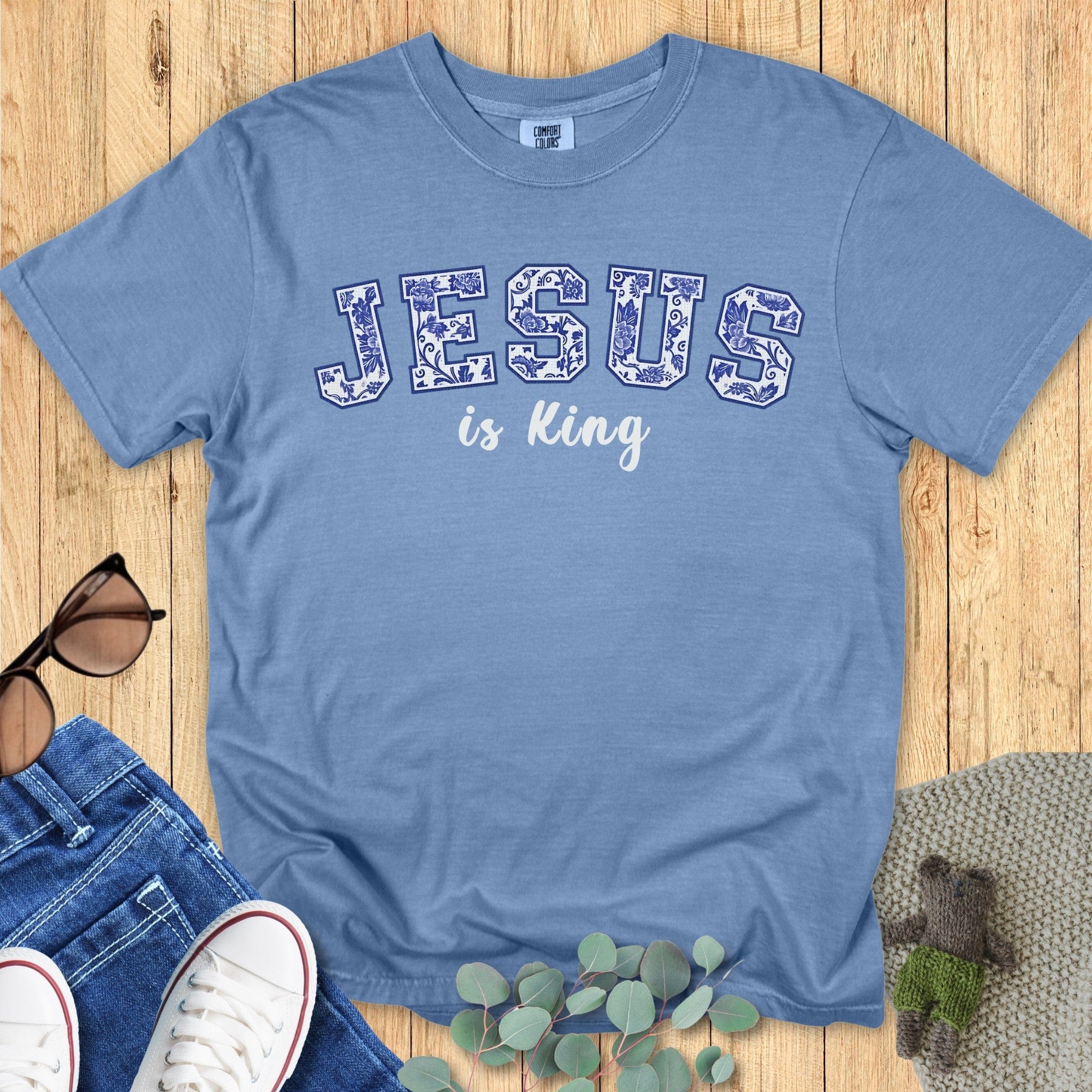 latlay of a Comfort Colors 1717 blue jean “Jesus is King” t-shirt featuring bold blue floral collegiate letters. A soft women’s scripture tee - faith-based apparel for women and modern Christian shirt, thoughtful Christian mom gift idea that celebrates faith in style.