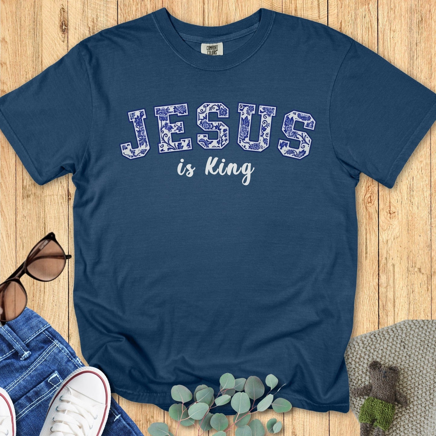 Flatlay of a Comfort Colors 1717 midnight “Jesus is King” t-shirt with intricate blue floral varsity-style letters. A premium women’s scripture tee - faith-based apparel for women and modern Christian shirt, meaningful Christian mom gift idea for worship and casual wear.