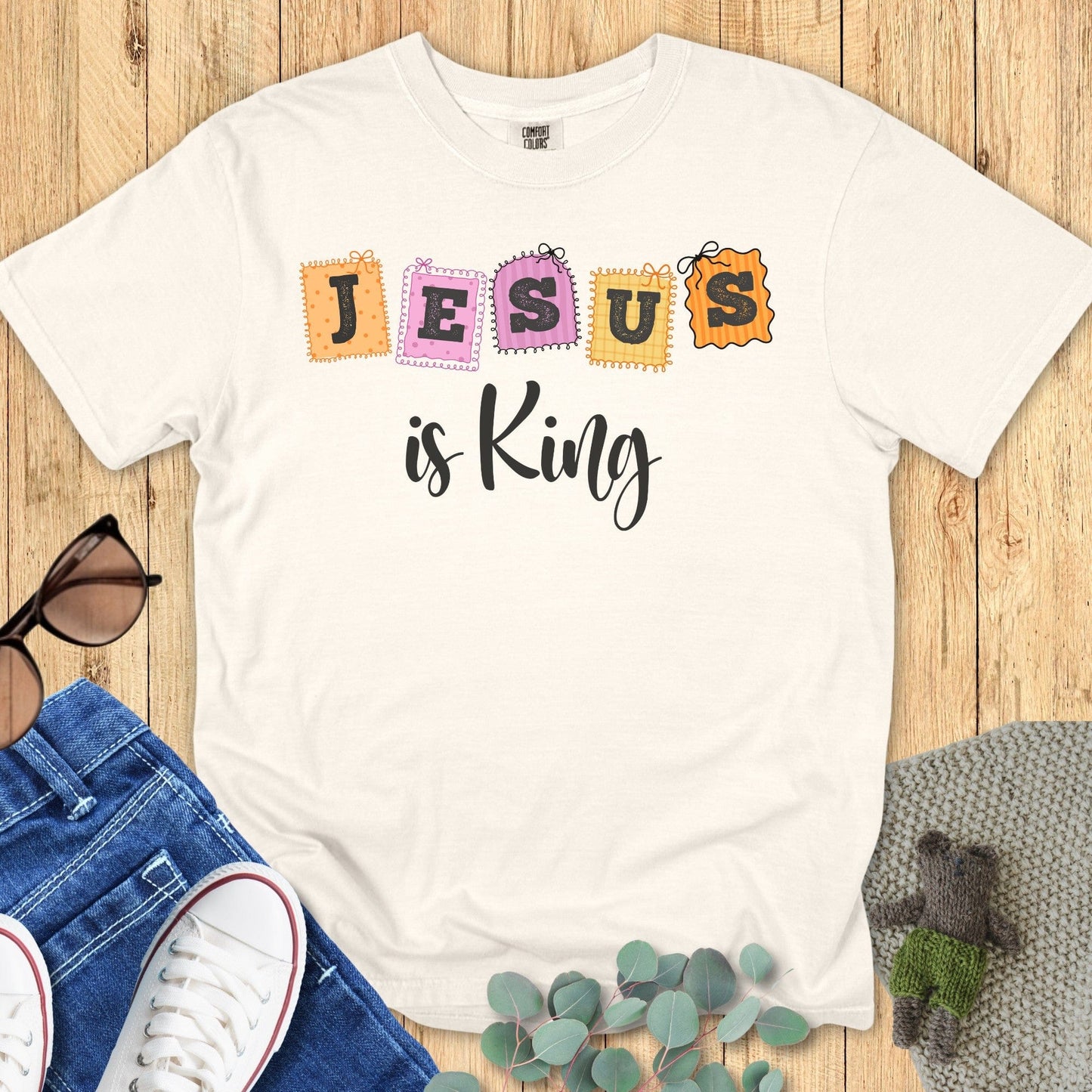 Flatlay of a Comfort Colors 1717 ivory “Jesus is King” t-shirt featuring pastel stitched block letters. A classic women’s scripture tee - faith-based apparel for women and modern Christian shirt, beautiful Christian mom gift idea for daily inspiration.