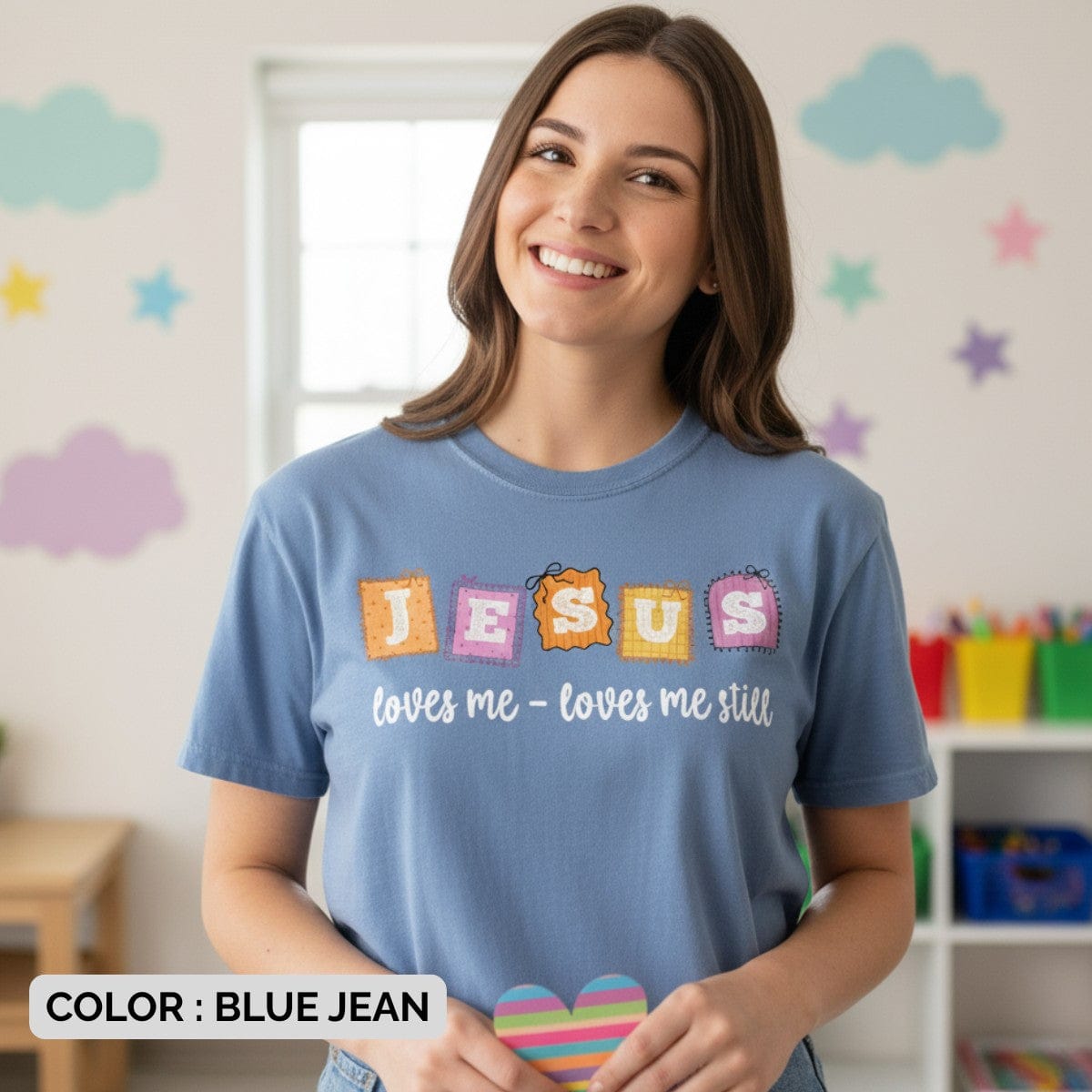 Smiling woman wearing a Comfort Colors 1717 blue jean “Jesus loves me – loves me still” t-shirt with colorful patch-style lettering. A joyful women’s scripture tee — faith-based apparel for women and modern Christian shirt, thoughtful Christian mom gift idea for everyday encouragement.