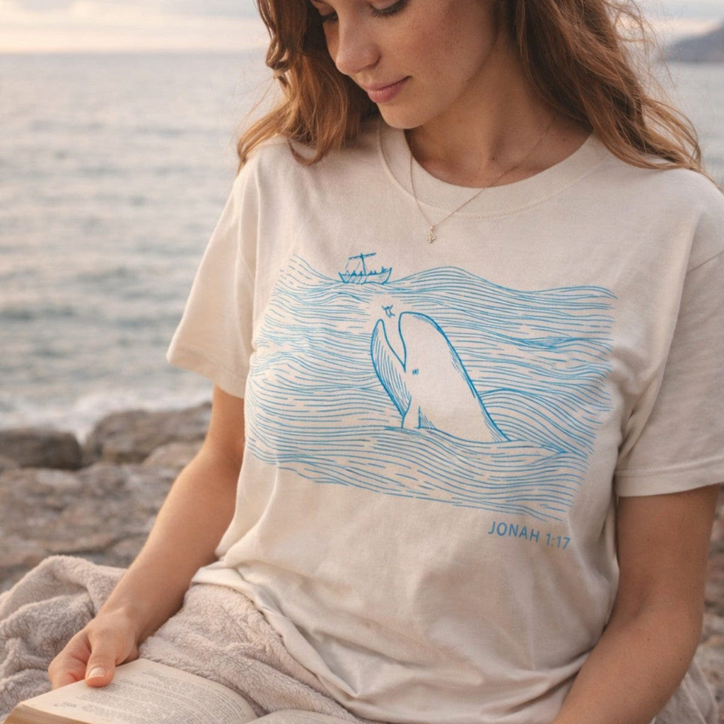 Woman reading a Bible by the sea wearing an Ivory Comfort Colors 1717 tshirt with a blue line drawing of a whale. A serene Christian women tee featuring Jonah 1:17 scripture, perfect for coastal US faith wear styles.