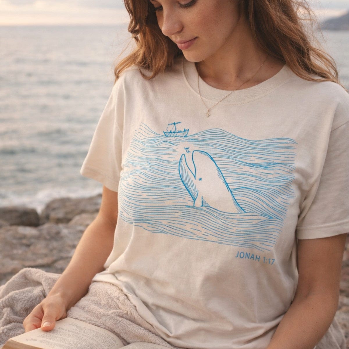 Woman reading a Bible by the sea wearing an Ivory Comfort Colors 1717 tshirt with a blue line drawing of a whale. A serene Christian women tee featuring Jonah 1:17 scripture, perfect for coastal US faith wear styles.