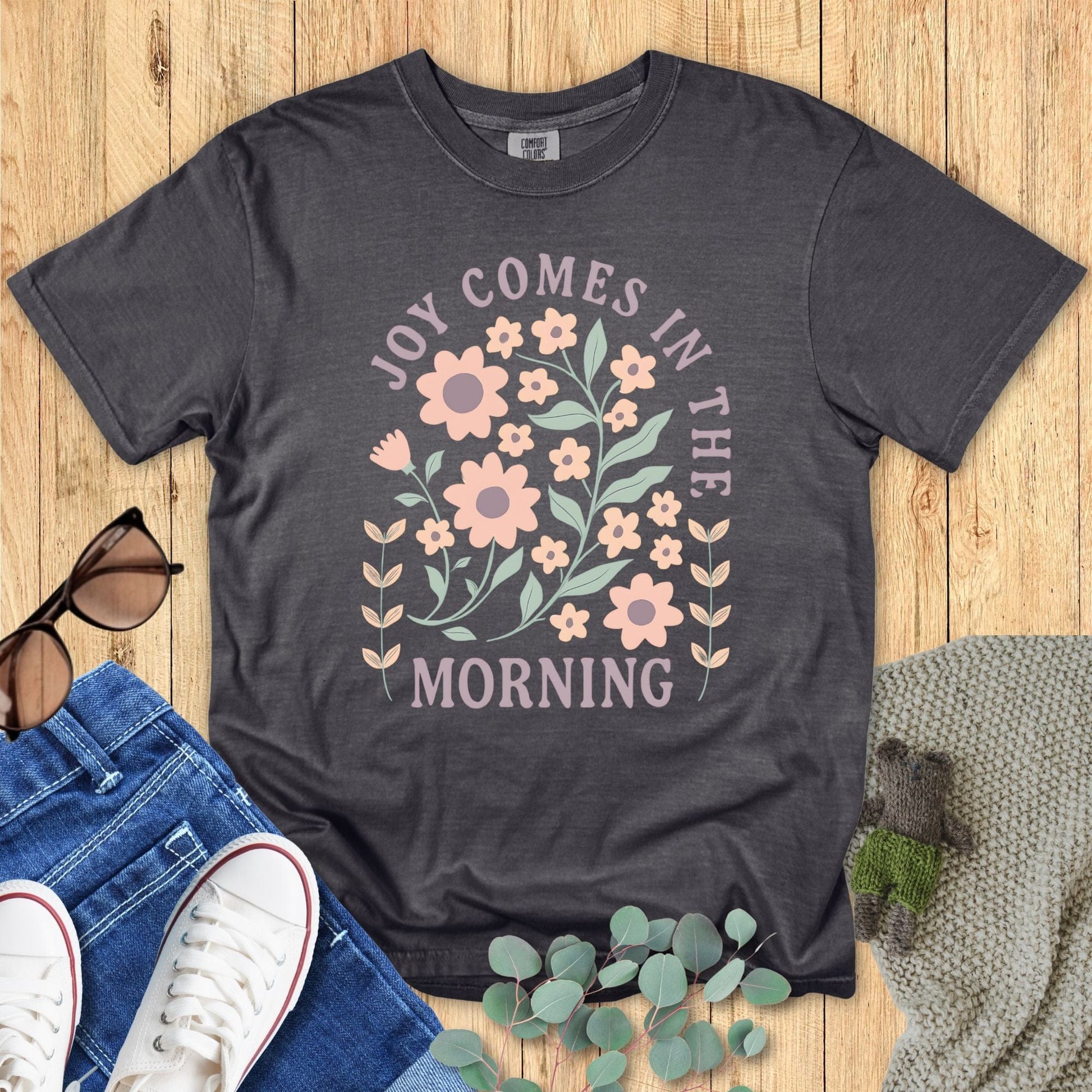 Comfort Colors Christian T-shirt in pepper gray with floral design and scripture quote 'Joy Comes in the Morning.' Inspirational faith-based graphic tee styled flat lay with jeans and sunglasses.