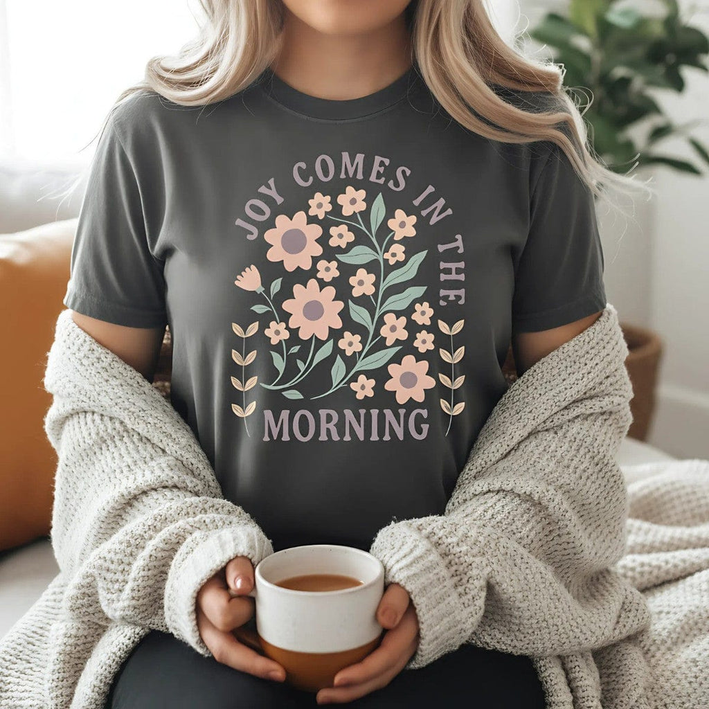 "Christian woman wearing a Comfort Colors Christian T-shirt in pepper gray with floral design and verse 'Joy Comes in the Morning.' Cozy faith-based outfit styled with cardigan and coffee cup indoors."