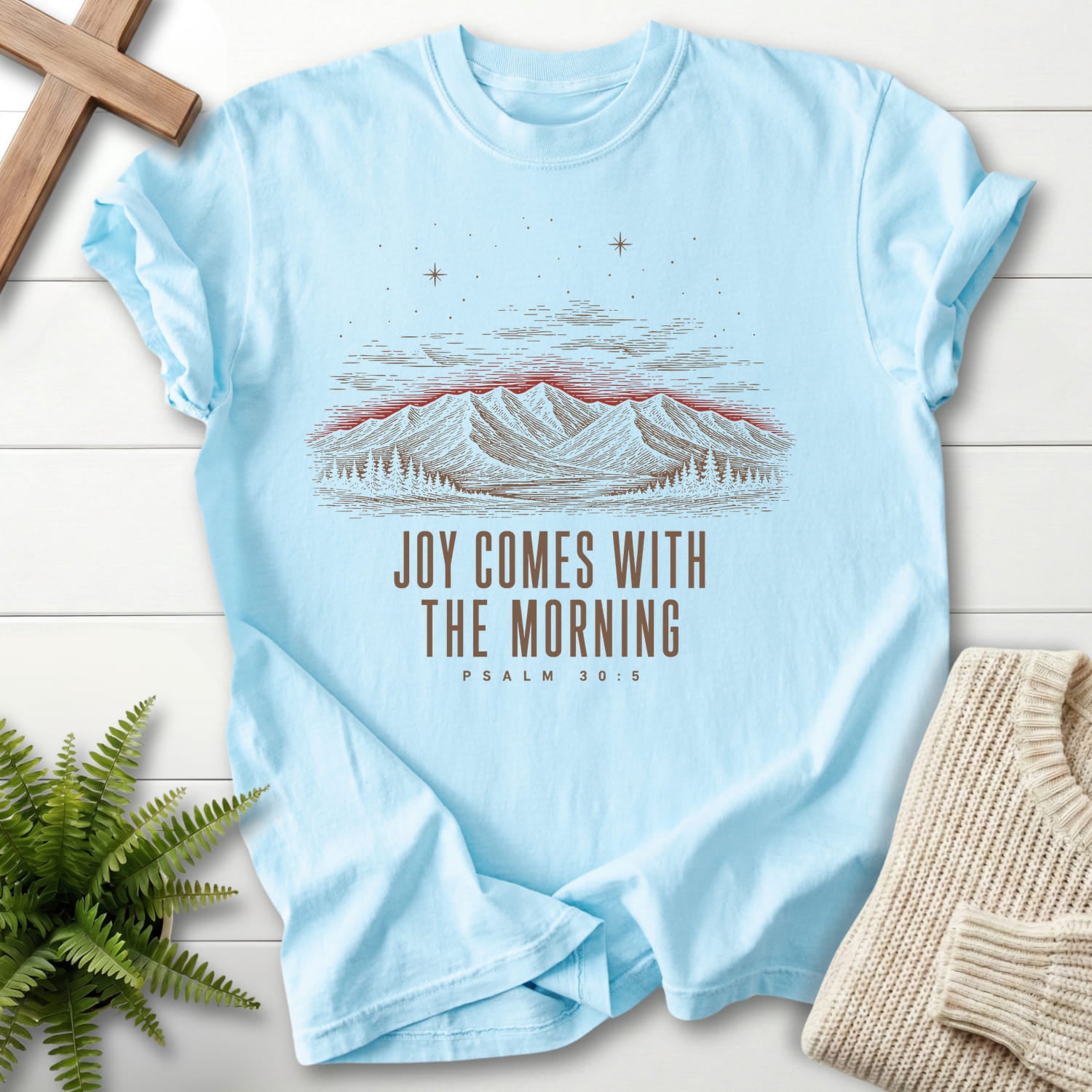 Flatlay of a Chambray Christian t-shirt for women with mountain graphic with "Joy Comes With the Morning Mountain T-Shirt" Psalm 30:5.