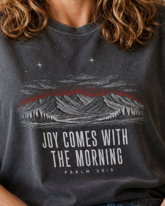 JOY Comes WITH - Pepper women's Christian tee, lifestyle photo in cozy reading nook - Engraved mountain range and pines under a starry sky - Psalm 30:5 - She Wears Faith