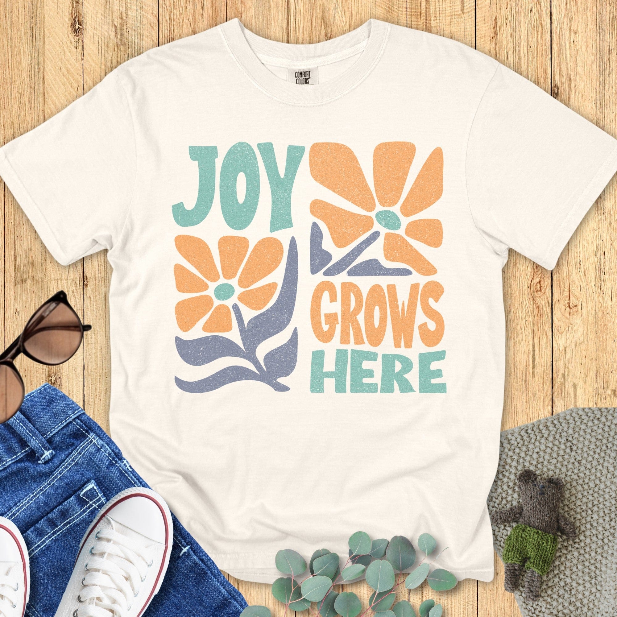 Flat lay of a Ivory T-shirt with the same “Joy Grows Here” floral design, styled on a wooden background alongside sunglasses, jeans, white sneakers, and greenery