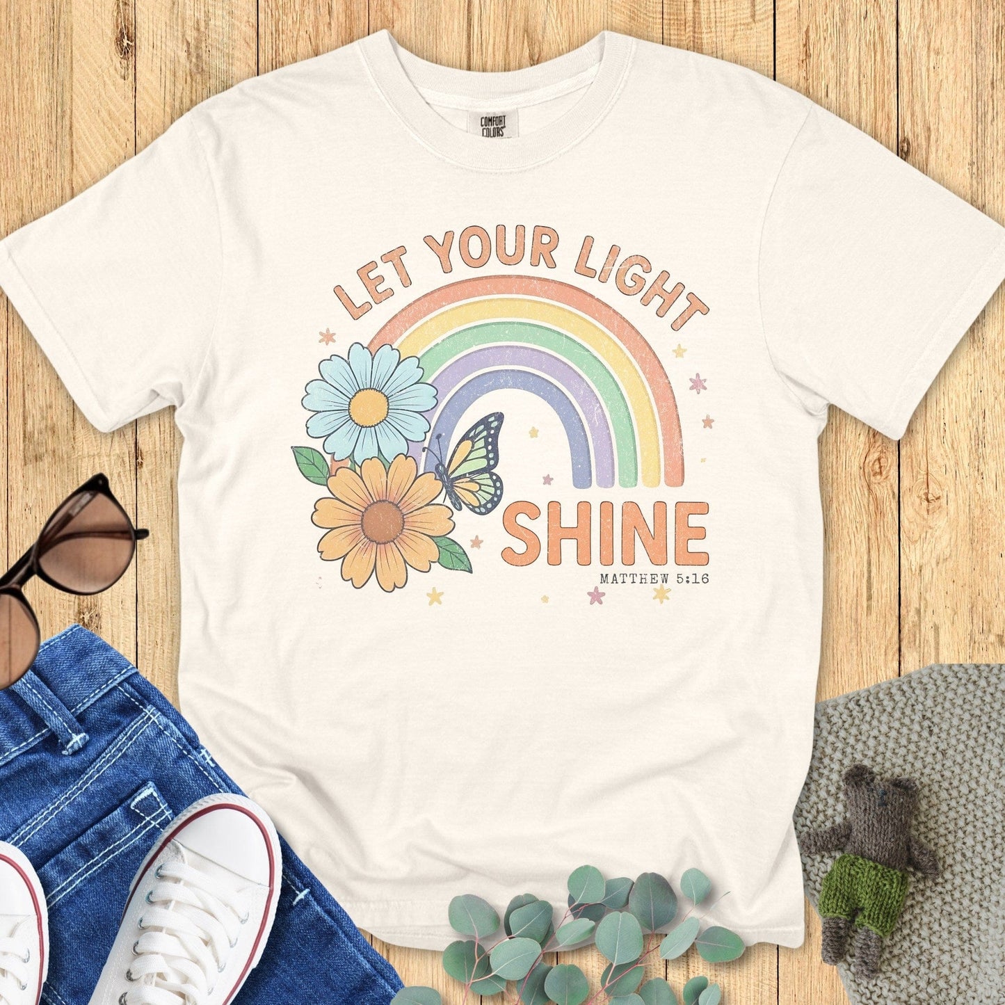 Flatlay of a Comfort Colors 1717 ivory “Let Your Light Shine” Matthew 5:16 t-shirt with retro rainbow, floral, and butterfly design. A bright women’s scripture tee - faith-based apparel for women and modern Christian shirt, perfect Christian mom gift idea that spreads joy and faith.