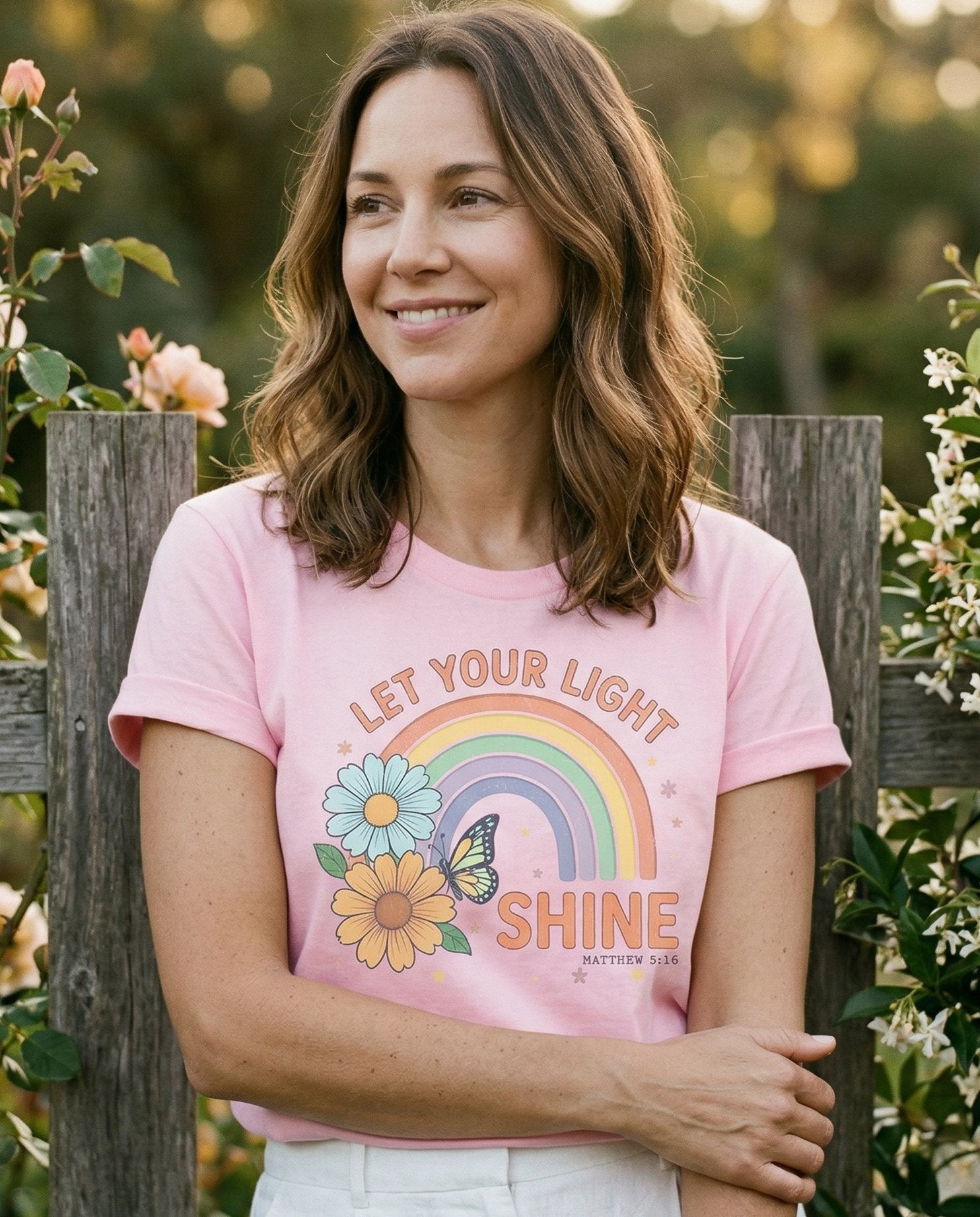 Let Your Light Shine – Blossom women's Christian tee, lifestyle photo in outdoor garden – Retro groovy design with a pastel rainbow arc – Matthew 5:16 – She Wears Faith