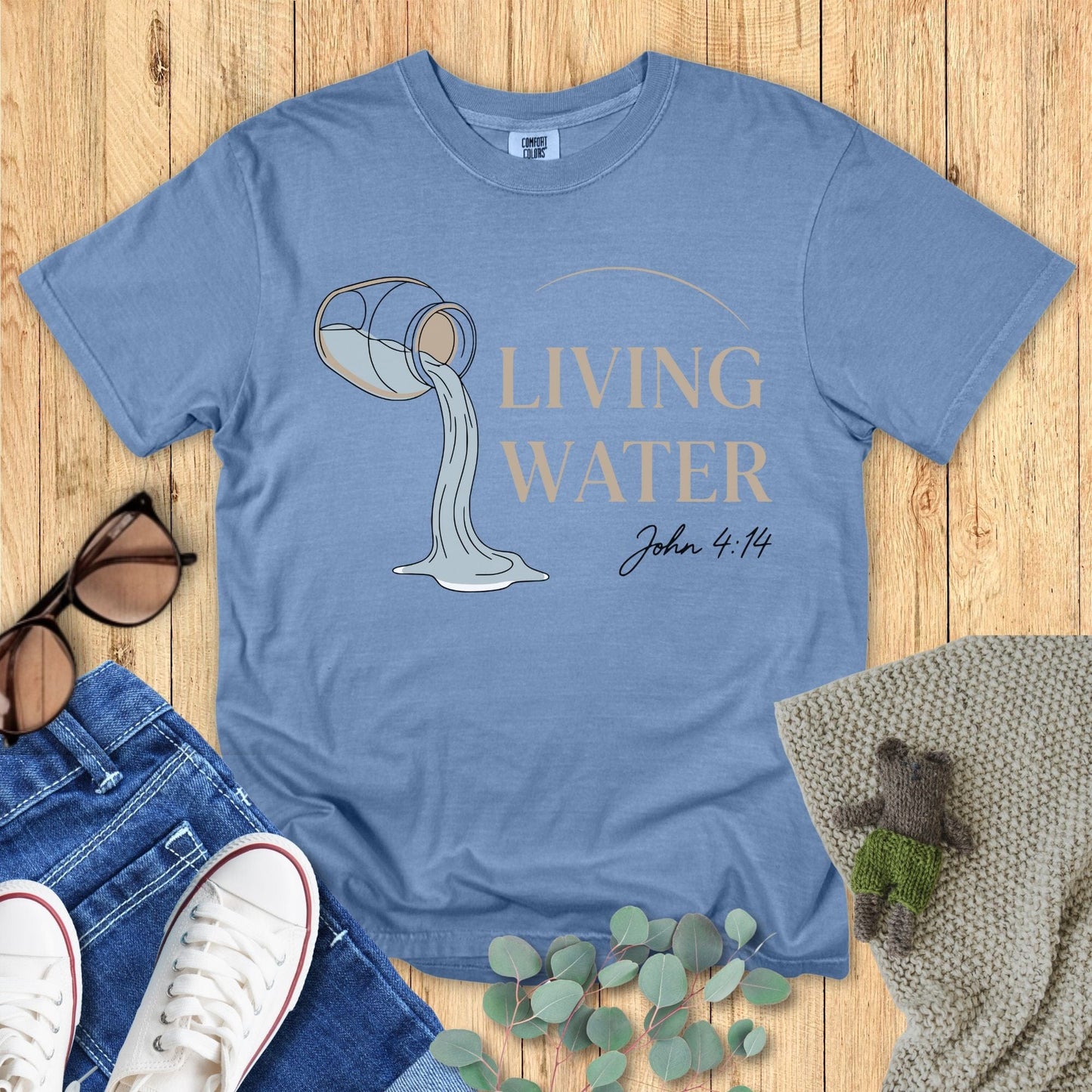 Flatlay of a Comfort Colors 1717 blue jean “Living Water – John 4:14” t-shirt with tan lettering and flowing water graphic. A calming women’s scripture tee — modern faith-based apparel for women and perfect Christian mom gift idea inspired by John 4:14.