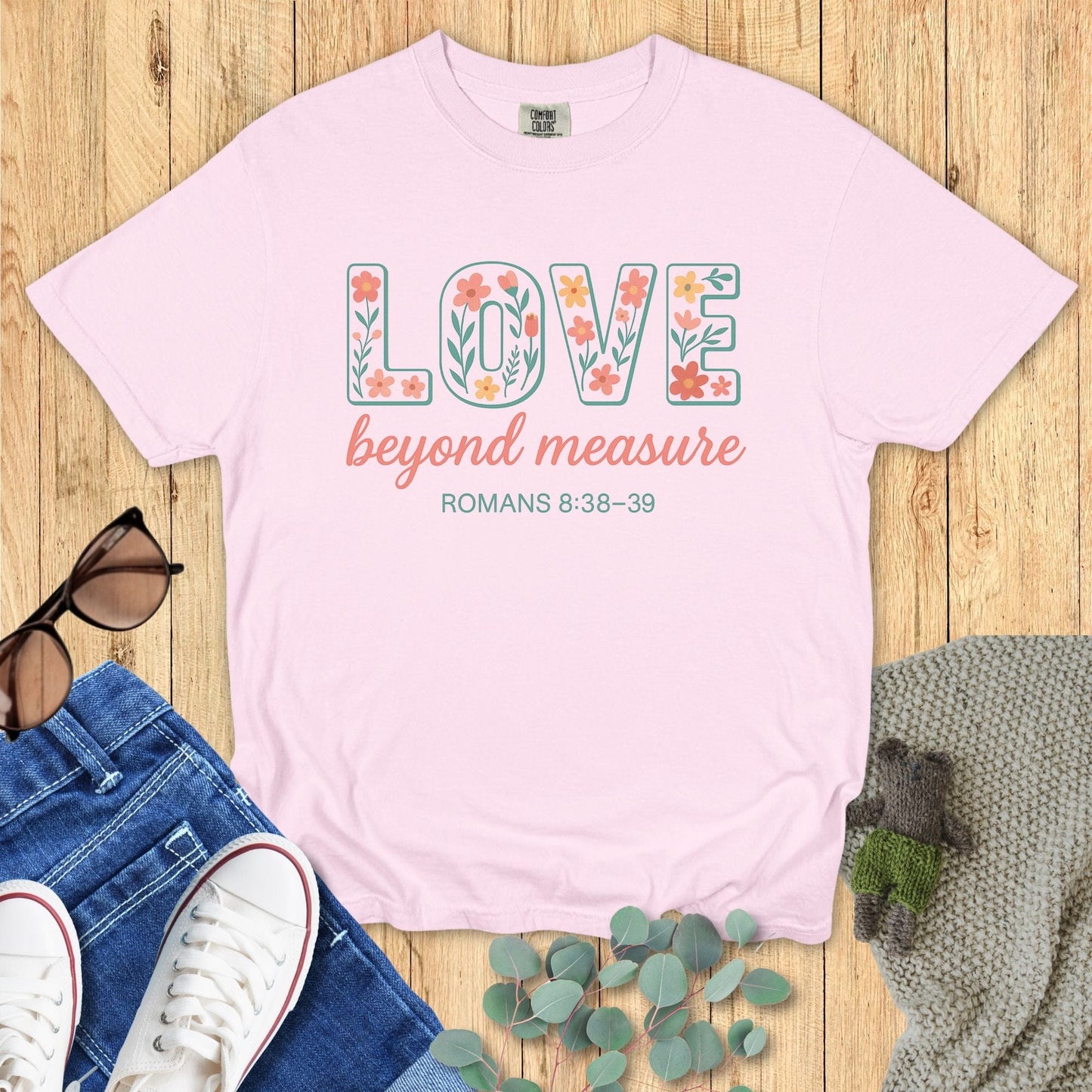 Blossom pink Comfort Colors Christian T-shirt with floral block text "LOVE beyond measure," and verse Romans 8:38–39. Faith-based inspirational tee flat lay with jeans, sneakers, and greenery on wood surface.