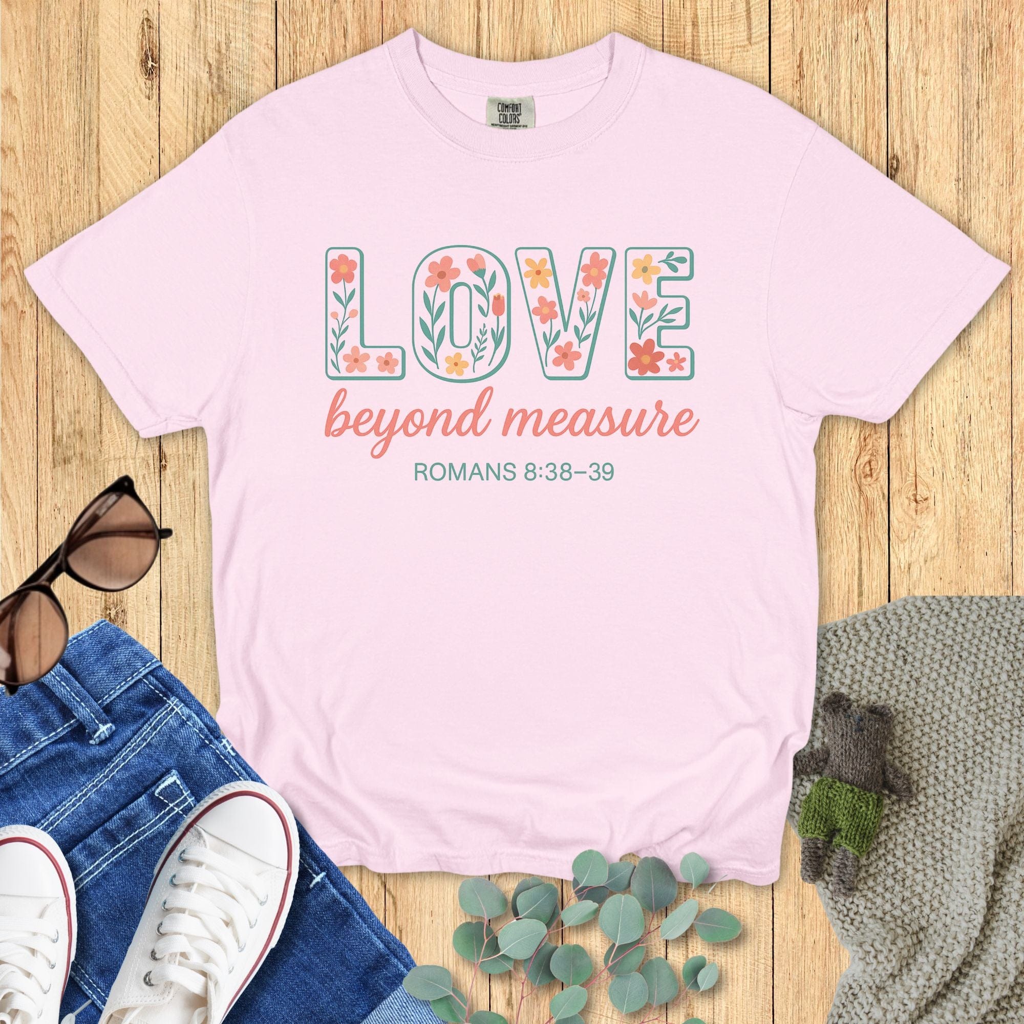 Blossom pink Comfort Colors Christian T-shirt with floral block text "LOVE beyond measure," and verse Romans 8:38–39. Faith-based inspirational tee flat lay with jeans, sneakers, and greenery on wood surface.