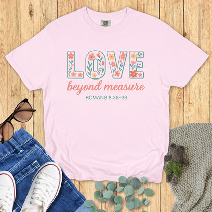 Blossom pink Comfort Colors Christian T-shirt with floral block text "LOVE beyond measure," and verse Romans 8:38–39. Faith-based inspirational tee flat lay with jeans, sneakers, and greenery on wood surface.