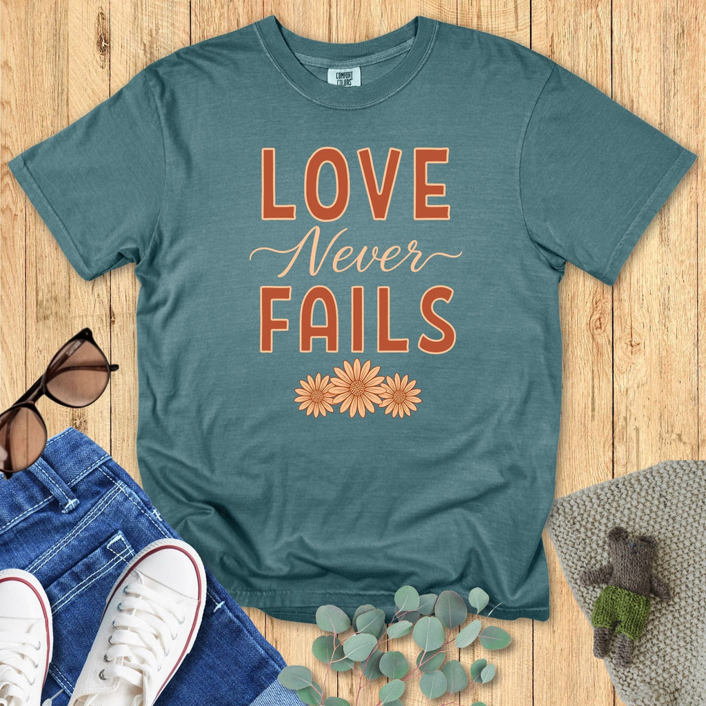 Comfort Colors 1717 blue spruce “Love Never Fails – 1 Corinthians 13:8” t-shirt featuring warm retro-style typography and daisy motifs. A stylish women’s scripture tee — vintage-inspired faith-based apparel for women and thoughtful Christian mom gift idea.