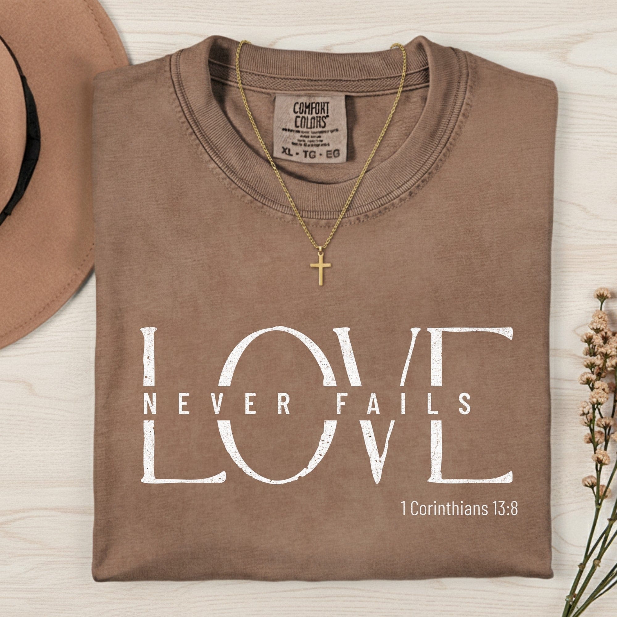 Folded Comfort Colors 1717 espresso “Love Never Fails” t-shirt paired with a gold cross necklace. A minimalist women’s scripture tee — faith-based apparel for women and modern Christian shirt inspired by 1 Corinthians 13:8, meaningful Christian mom gift idea.